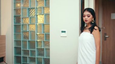 Beyaz bir havlu ve zarif mücevherler içinde büyüleyici bir kadın modern bir otel odasında duruyor, düşünceli bir şekilde yan tarafa bakıyor..