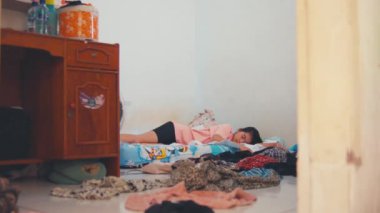 Geniş açıda dağınık bir oda, yere dağılmış giysiler ve yatağında uyuyan genç bir Asyalı kadın görülüyor. Düzensiz ve muhtemelen stresli bir yaşam ortamını veya yoğun bir çalışma dönemini vurguluyor..