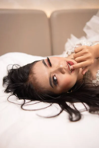 an Asian woman with black hair biting her fingers with a horny expression while sleeping on a white bed at night