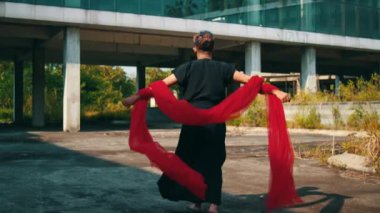 Asyalı bir adam, gün içinde terk edilmiş bir binada rüzgarda uçan kırmızı bir eşarpla koşup dans ediyor.