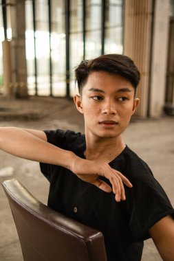 Eski bir binada siyah pantolon giyerken kahverengi deri koltukta poz veren Asyalı bir adam.