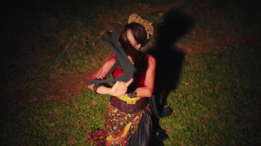 Asyalı bir kadın yeşil bir çimende oturmuş geceleri ormanda elleri ve kırmızı bir eşarpla dans ediyor.