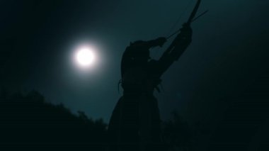 Siluetli bir kadın okçu gece yayını uçurumdan çekerken ayakta duruyor.