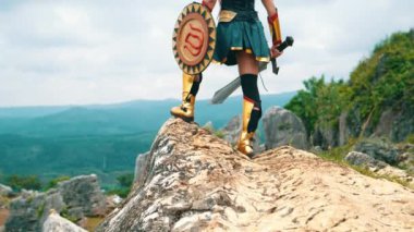 Bir kadın savaşçı elinde kılıç ve altın kalkanla gündüz vakti bir uçurumun kenarında cesurca duruyor.