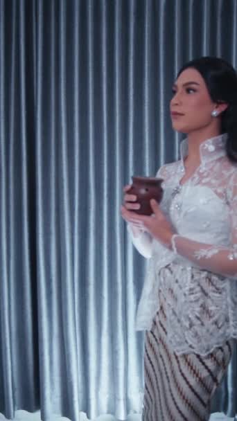 穿着传统服装的优雅女人 拿着杯子 带着若有所思的表情 背对着窗帘 — 图库视频影像