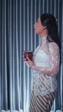 Geleneksel giyinmiş zarif bir kadın elinde fincanla, sakin bir ifadeyle perde arkasında duruyor..