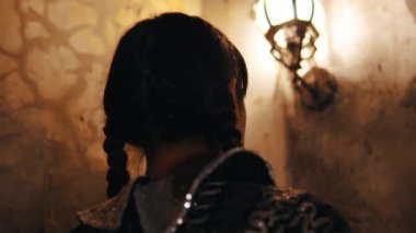 Saçları örülmüş bir kişinin silüeti dokulu bir duvara karşı, gecenin karanlığında sıcak sconce ışıklarıyla aydınlatılıyor.