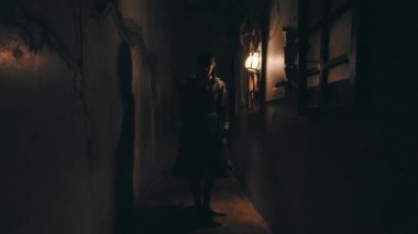 Karanlık, dar bir sokakta ya da koridorda siluet içinde duran bir kadının tam görüntüsü. Tek bir sıcak duvar feneri ürkütücü, atmosferik aydınlatma sağlar ve uzun gölgeler bırakır. Gizem ve belirsizlik.