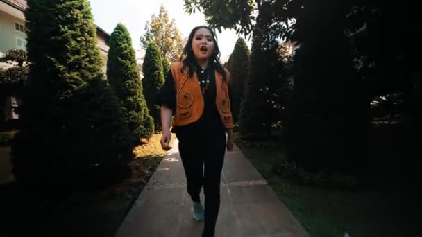 バックグラウンドの自然な日光と緑で屋外に出る黄色いベストのスタイリッシュな若い女性 — ストック動画