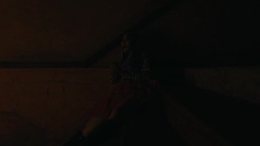 Karamsar bir kadın portresi, dramatik ışıklandırma ve canlı kırmızı etekle merdivenlerin altında oturuyor, gece boyunca tefekkür ediyor ya da yalnızlık çekiyor.