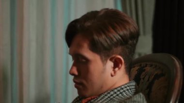 Sandalyede oturan bir erkek tasarımcı, mücevher üzerinde çalıştıktan sonra bitkin görünüyor. Elinde gümüş bir taç var, işini düşünüyor ya da mükemmeliyetçiliğin ağırlığını hissediyor..