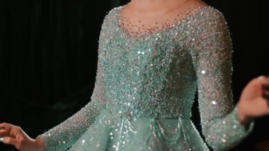 Bir mankenin ellerini dinlendirirken ve ışıl ışıl elbisesinde zarifçe hareket ederken gösteren detaylı bir fotoğraf. Video, kumaşın dokusunu ve manikürlü tırnakların karanlık bir ortamda olduğunu vurguluyor..