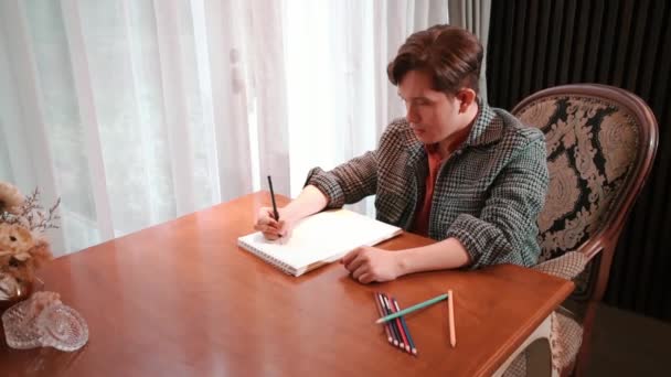 穿着粗花呢夹克的男人在一张木制桌子旁的笔记本上写字 桌上有铅笔和一个花瓶 窗前有巨大的窗帘 — 图库视频影像