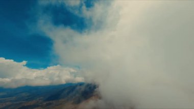 Dağlık arazi üzerinde mavi gökyüzü ile dramatik bulutlar, gün ışığında fırtınalı ve açık hava arasındaki zıtlığı betimliyor.