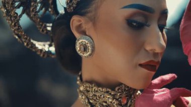 Karmaşık mücevherleri ve makyajı olan, kırmızı çiçekle poz veren zarif bir kadın, gün ışığında güzellik ve lüks sergiliyor.