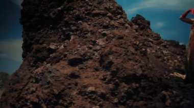 Mitolojik kostümlü kadın gün ışığında dramatik bir gökyüzünün altında kayalar arasında poz veriyor.