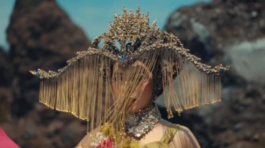 Arkadan çekilen bir kare altın bir başlık ve renkli bir elbisenin arkasını ayrıntılı bir şekilde gösteriyor. Şekil sert, kayalık bir çöle bakıyor, bir yolculuk ya da tefekkür izlenimi veriyor..