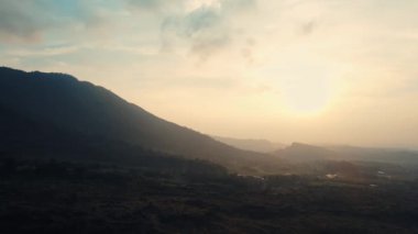 Gün ışığında yumuşak ışık ve katmanlı tepelerle sakin bir dağ manzarasının üzerinde puslu bir gün doğumu.