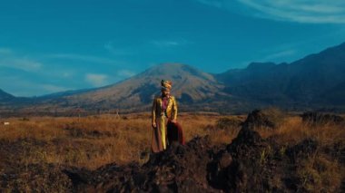Geleneksel elbiseli kadın gün batımında bir dağın önünde duruyor.