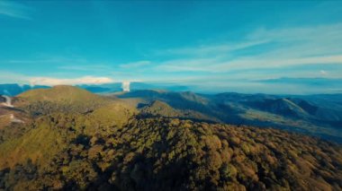 Berrak mavi gökyüzünün altında, gün ışığında ince bulutlu, yemyeşil bir dağ sırasının havadan görünüşü.