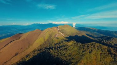 Gün ışığında açık mavi gökyüzünün altında yemyeşil bir dağ sırtı görünüyor.