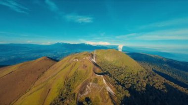 Arkasında açık mavi gökyüzü olan yemyeşil bir dağ sırasının havadan görünüşü.