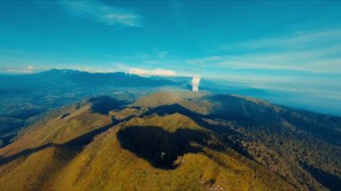 Açık mavi gökyüzü ve muhtemel bir orman yangını veya endüstriyel kaynaktan gelen uzak duman bulutlarıyla dağlık bir arazinin havadan görünüşü.