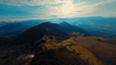Gün batımında bulutlu bir gökyüzünün altında yuvarlanan tepelerin hava görüntüsü, gün ışığında doğal manzaranın dingin güzelliğini gözler önüne seriyor.