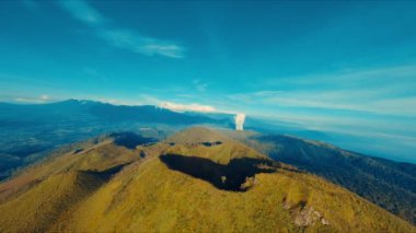 Bereketli yeşillikle dağlık arazinin havadan görünüşü ve gün ışığında açık mavi gökyüzüne yükselen uzak bir duman bulutu.