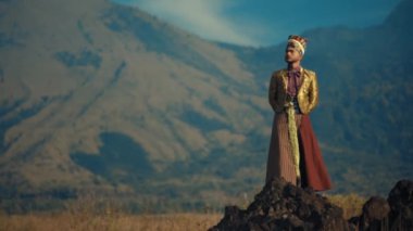 Geleneksel kıyafetli bir insan gün batımında dağlık bir zemin önünde duruyor.