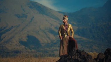 Geleneksel giyinmiş bir kadın gün batımında dağ manzarasının önünde duruyor.