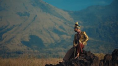 Geleneksel giysili adam dağ manzarasının önünde duruyor, gün ışığında doğayı düşünüyor.