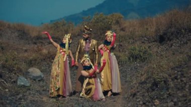 Geleneksel kostümlü üç kişi gün ışığında dağlık bir zemin üzerinde kültürel bir dans gösterisi yapıyorlar.