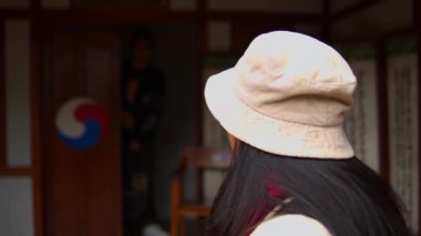 Ön planda şapkalı bir kadın, sabahları Kore bayrağıyla geleneksel bir kapıda dikilen birine bakıyor.