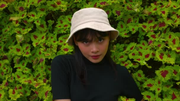 一个戴着白帽的年轻女子面对着红色花朵的生机勃勃的绿叶背景时的画像 — 图库视频影像