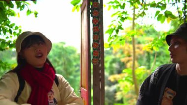 Sıradan giyinen iki turist, geleneksel ahşap bir yapının içinde sabahları arka planda yeşillik tartışıyor.