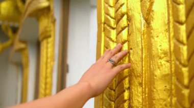 Genç bir kadının eline yakın plan, yüzük takıyor, geleneksel bir Asya tapınağındaki süslü altın bir sütunun karmaşık, dokulu yüzeyine nazikçe dokunuyor ve kayıyor..