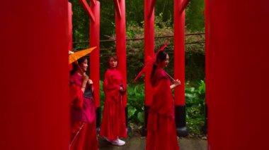 Genç bir turist çift, geleneksel kırmızı elbiseli (kimono / yukata) bir grup kadının yanından geçer. Kadınlar kırmızı sütunlu Japon tarzı bir tapınakta renkli kağıt şemsiyelerle poz veriyorlar..