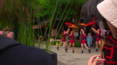 Bir çiftin diziyi izleyip eğlenirken omuz üstünden çekimleri. Kadın alkışlar ve erkek kültürel bir köyde kağıt şemsiyelerle dans eden kırmızı elbiseli bir grup kadın sanatçıya onay verir..