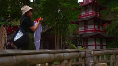 Bir köprüde duran şapkalı ve sırt çantalı gezgin, sabah yemyeşil bir parktaki geleneksel kırmızı pagoda 'ya hayran.