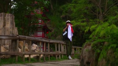 Sırt çantalı gezgin, yemyeşil bir parktaki ahşap bir köprüde duruyor ve sabahları geleneksel kırmızı pagoda 'ya hayran kalıyor.