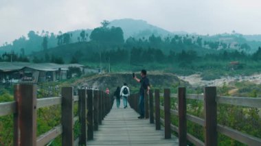 Genç bir adam uzun ahşap bir köprüde duruyor, elinde akıllı telefonuyla volkanik bir platonun sisli manzarasıyla çevrili bir selfie çekiyor..