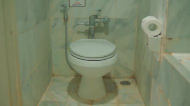 Temel banyo içi beyaz seramik tuvalet, duvara monte edilmiş lavabo ve otel odasının içinde tuvalet kağıdı tutacağı var.