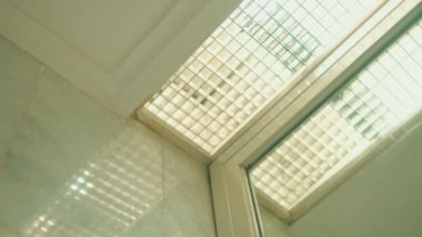 Parlak güneş ışığı cam bloktan içeri akıyor, gün ışığında fayanslı bir zemine desenli bir gölge bırakıyor.