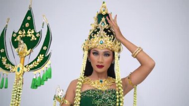 Süslü kostümlü geleneksel Endonezyalı dansçı stüdyoda beyaz arka planda zarif el hareketleriyle performans sergiliyor.