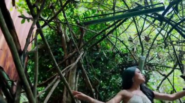 Düşük açılı bir çekimde bir kadın elini bir dala uzatıyor ve yemyeşil bir ortamda meraklı ve sakin bir ifadeyle yukarı bakıyor..