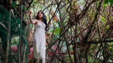 Beyaz elbiseli, Asyalı bir kadının, sarmaşık ve dallardan oluşan yoğun bir yapıda yürürken, etrafa bakındığı orta boy bir fotoğraf..