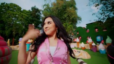 Dans eden ve saçlarını savuran Asyalı genç bir kadının orta boy yakın çekimi. Arka plandaki dansçıları tema parkındaki ahşap köprüde gösteri yaparken kameraya gülümsüyor..