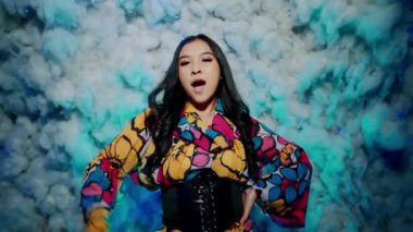 Kameraya bakıp poz veren kendinden emin genç bir kadının portresi. Renkli bir çiçek bluzu ve siyah bir korse giyiyor. Mavi ışıklı yapay bulutların önünde duruyor..