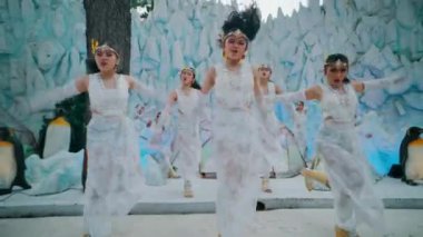 A group of beautiful Asian dancers in white lace costumes performing an energetic choreography on a stage decorated like an ice castle with snow and penguins. High energy performance.